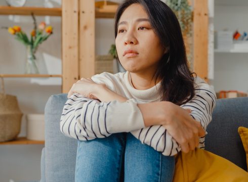 Thoughtful Asia lady sit embracing knees at sofa in living room at house look outside with feel lonely, Sad depressed teenager spend time alone stay at home, Social distance, Coronavirus Quarantine.